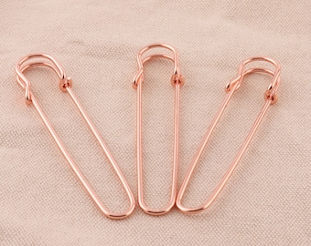 May include: Three rose gold safety pins on a light brown fabric background.