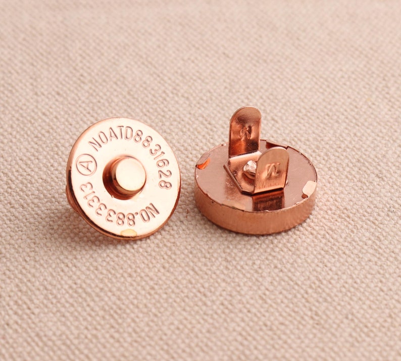 May include: Two copper-coloured magnetic snaps, one with a round base and the other with a base that has a metal loop. The round base has the text "NOATD8831628 E1633 88'ON" engraved on it.