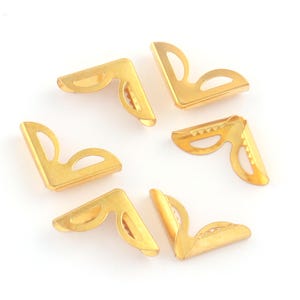 May include: Six gold-colored metal corner protectors, arranged on a white surface. Each L-shaped piece has a decorative cutout and a folded edge for securing to a corner. These are often used in scrapbooking and crafting.