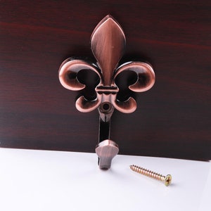 May include: A copper-colored metal hook with a fleur-de-lis design. The hook is attached to a wall with a screw.