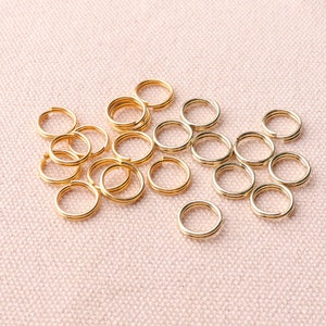 May include: A close-up of a group of gold jump rings. The rings are arranged in a random pattern on a pink fabric background.