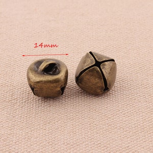 May include: Two antique bronze-colored bells, each approximately 0.55 inches (14mm) in diameter. One bell has a loop for hanging, while the other has a cross-shaped design. The bells are likely for crafts or decorative purposes.