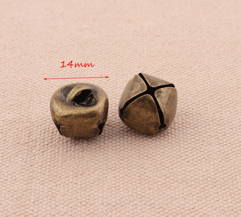 May include: Two antique bronze-colored bells, each approximately 0.55 inches (14mm) in diameter. One bell has a loop for hanging, while the other has a cross-shaped design. The bells are likely for crafts or decorative purposes.