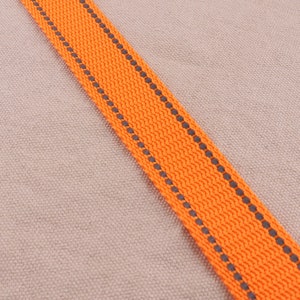 May include: Orange and gray woven ribbon with a reflective strip. The ribbon is on a beige background.