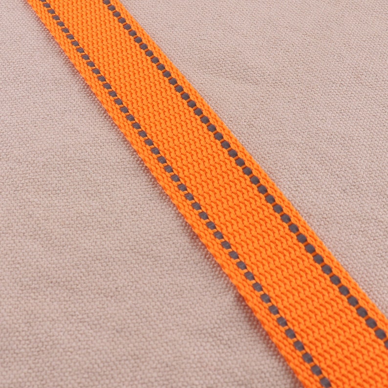 May include: Orange and gray woven ribbon with a reflective strip. The ribbon is on a beige background.