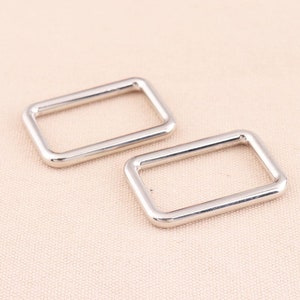 May include: Two silver metal rectangular rings, also known as D-rings, are shown on a light beige fabric surface.