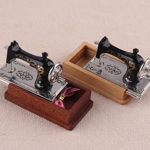 May include: Two miniature black sewing machines with gold accents are displayed in wooden boxes. The sewing machines have a silver base and a floral design. The boxes are made of light brown wood.