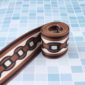 May include: Brown and white patterned fabric ribbon with a chain link design. The ribbon is rolled up and the chain link design is visible on the front and back of the ribbon.