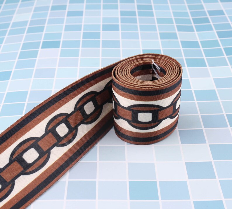 May include: Brown and white patterned fabric ribbon with a chain link design. The ribbon is rolled up and the chain link design is visible on the front and back of the ribbon.