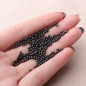May include: A large pile of small, round, black beads. The beads are smooth and shiny, and they are scattered on a hand.