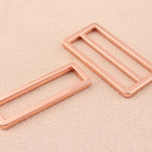 May include: Two rose gold metal rectangular frame buckles. The larger buckle has two horizontal bars inside the frame. The smaller buckle has one horizontal bar inside the frame.