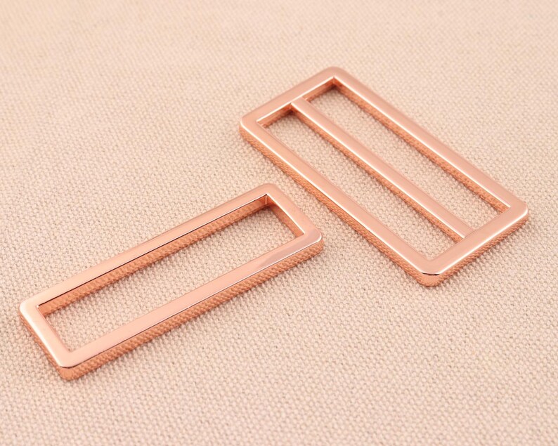 May include: Two rose gold metal rectangular frame buckles. The larger buckle has two horizontal bars inside the frame. The smaller buckle has one horizontal bar inside the frame.