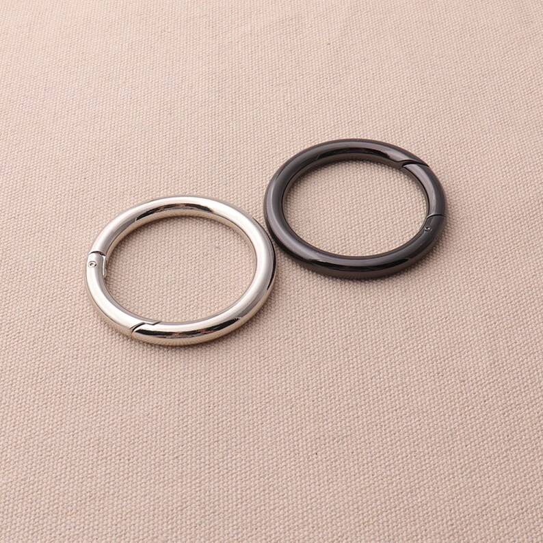 May include: Two metal rings, one silver and one black, with a snap closure. The rings are on a beige fabric background.