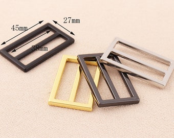 Könnte beinhalten: Vier Metall-Schieber-Schnallen in Gold, Silber und Schwarz. Die Schnallen sind rechteckig mit zwei parallelen Linien in der Mitte. Die größte Schnalle misst 11,4 cm x 3,8 cm.