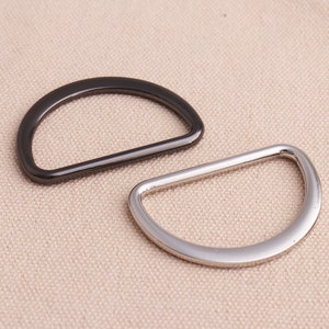 May include: Two metal D-rings, one black and one silver, on a beige fabric background.