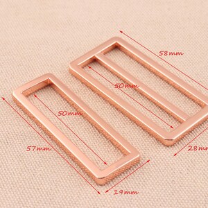 May include: Two rose gold metal rectangular belt buckle slides. The larger slide measures 5.8 inches by 2.8 inches and the smaller slide measures 5.7 inches by 1.9 inches.