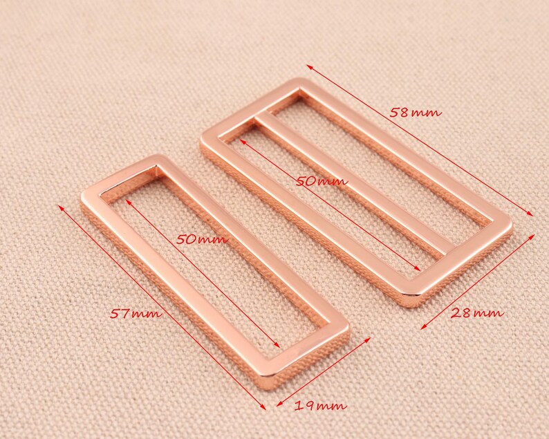 May include: Two rose gold metal rectangular belt buckle slides. The larger slide measures 5.8 inches by 2.8 inches and the smaller slide measures 5.7 inches by 1.9 inches.