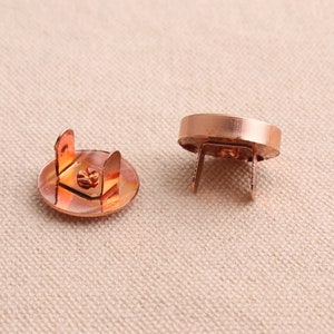 May include: Two copper magnetic snap closures. The closures are round with a flat top and a raised lip around the edge. The bottom of the closures has two prongs that extend out.
