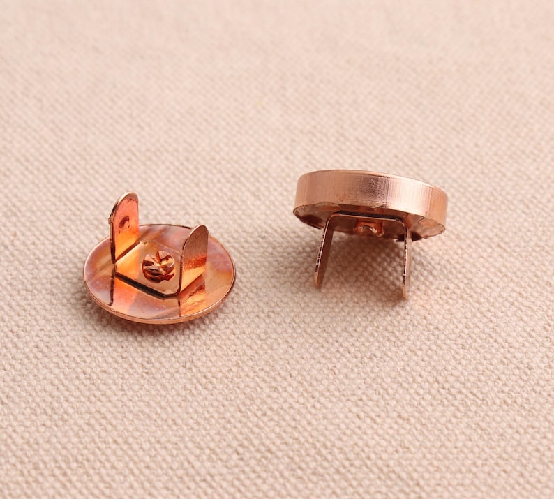 May include: Two copper magnetic snap closures. The closures are round with a flat top and a raised lip around the edge. The bottom of the closures has two prongs that extend out.