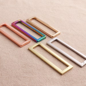 Könnte beinhalten: Fünf rechteckige Metallringe in verschiedenen Farben: Roségold, Regenbogen, Gold, Silber und Messing. Die Ringe sind in einer Reihe auf einer hellbraunen Stoffoberfläche angeordnet.