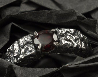Può includere: Un anello d'argento con una fascia testurizzata e una pietra preziosa rosso scuro. L'anello è in una scatola di velluto nero.