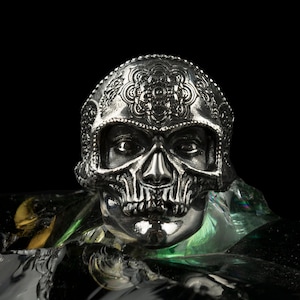 May include: A silver skull ring with intricate detailing on the forehead and around the eyes. The ring is set against a dark background, with a reflective surface below. The skull's features are clearly defined.