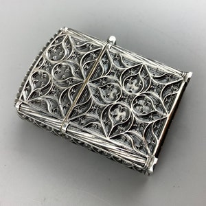May include: Silver lighter with an intricate, ornate design. The design features a repeating pattern of leaves and flowers.