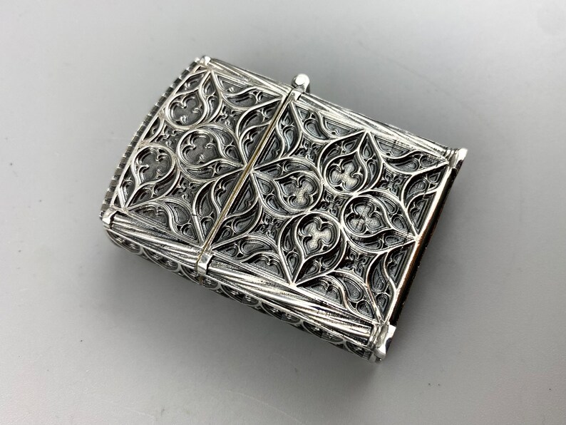 May include: Silver lighter with an intricate, ornate design. The design features a repeating pattern of leaves and flowers.