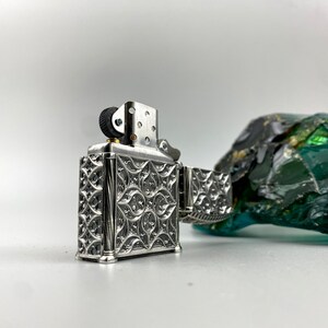 May include: A silver lighter with an intricate floral design. The lighter has a black wheel and a silver hinge.