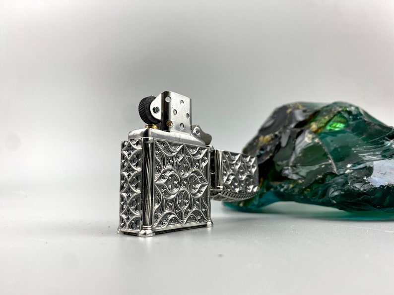 May include: A silver lighter with an intricate floral design. The lighter has a black wheel and a silver hinge.