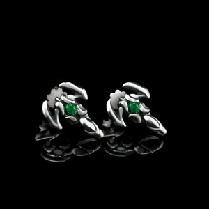 May include: A pair of silver earrings with a scorpion design and a green gemstone in the center of each scorpion.