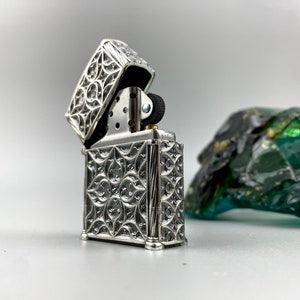 May include: A silver lighter with an intricate, ornate design. The lighter is open, revealing the flint and wick.