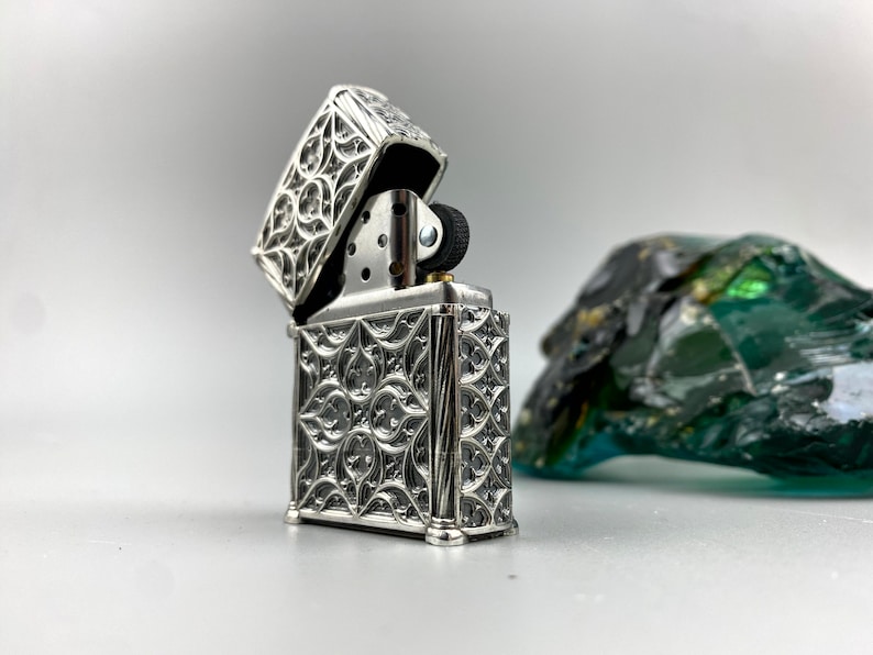 May include: A silver lighter with an intricate, ornate design. The lighter is open, revealing the flint and wick.