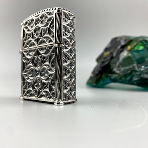 May include: A silver lighter with an intricate, ornate design. The lighter has a rectangular shape and a detailed, geometric pattern.