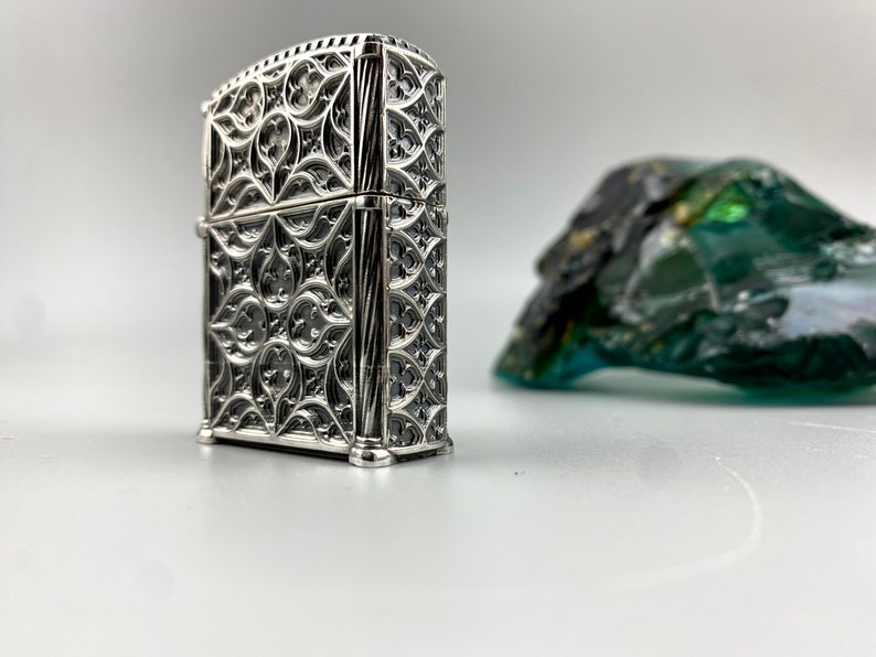 May include: A silver lighter with an intricate, ornate design. The lighter has a rectangular shape and a detailed, geometric pattern.