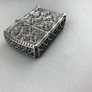 May include: A silver lighter with an intricate, ornate design. The lighter is closed and has a detailed, filigree pattern.