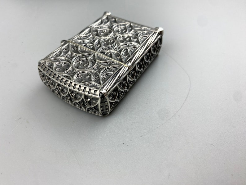 May include: A silver lighter with an intricate, ornate design. The lighter is closed and has a detailed, filigree pattern.