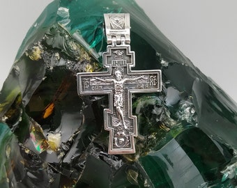May include: A silver cross pendant with intricate details and the inscription "Слава ХС" on the front. The cross is resting on a green, textured surface.