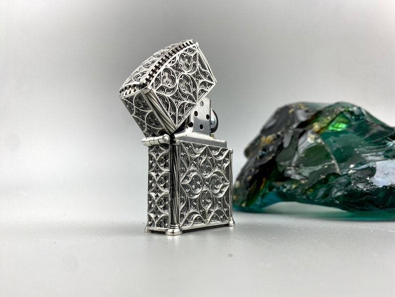 May include: A silver lighter with an intricate, ornate design. The lighter is open, revealing the flint and wick inside. The lighter is sitting on a white surface.