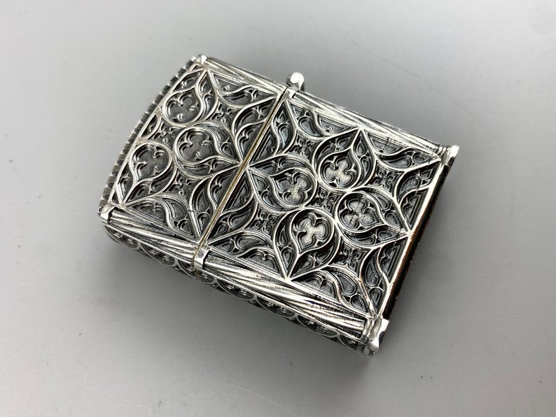 May include: Silver lighter with an intricate, ornate design. The design features a repeating pattern of leaves and flowers.