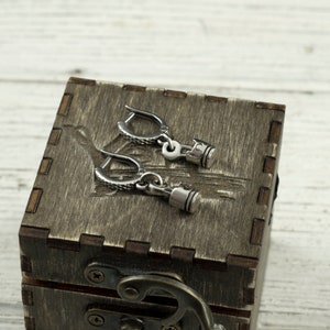 Puede incluir: Un par de pendientes de plata con un diseño de pistón. Los pendientes están en una caja de madera con un cierre.