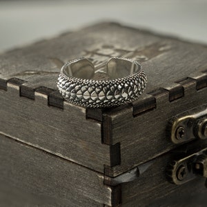 May include: A silver ring with a textured, scaly design sits on top of a wooden box with a brass latch.