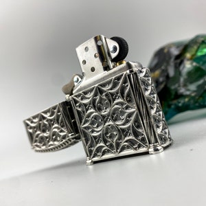 May include: A silver lighter with an intricate, ornate design. The lighter is open, revealing the inner mechanism. The lighter has a black wheel on the top.