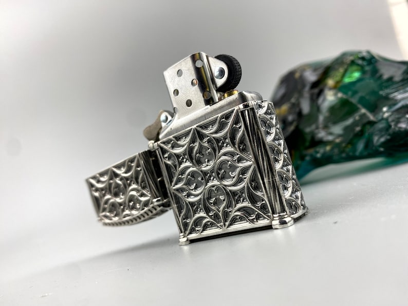 May include: A silver lighter with an intricate, ornate design. The lighter is open, revealing the inner mechanism. The lighter has a black wheel on the top.
