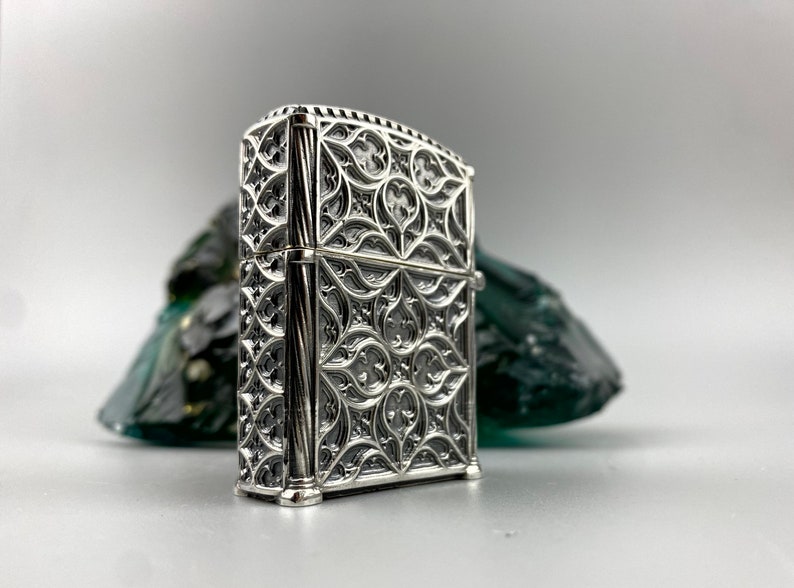 May include: A silver lighter with an intricate, ornate design. The design features a repeating pattern of geometric shapes and curves.