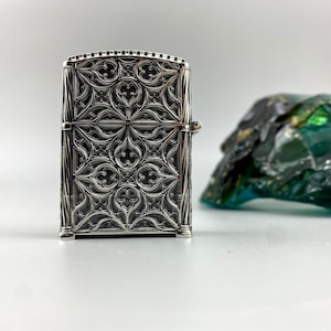 May include: Silver lighter with an intricate, geometric design. The design features a repeating pattern of arches and points, reminiscent of Gothic architecture.