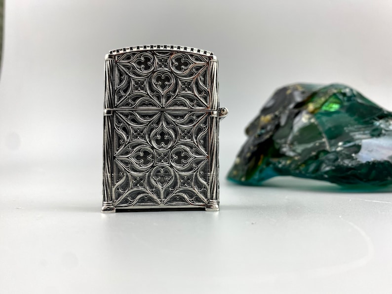 May include: Silver lighter with an intricate, geometric design. The design features a repeating pattern of arches and points, reminiscent of Gothic architecture.