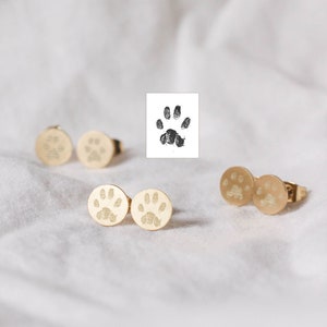 Op de afbeelding: Een paar gouden oorbellen met een pootprint ontwerp. De oorbellen liggen op een wit oppervlak.