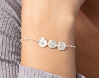 May include: A silver-toned chain bracelet featuring three small, circular charms, each engraved with a distinct paw print design. The delicate metal chain connects the charms in a horizontal row, creating a simple and refined piece of jewellery.