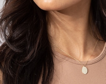 Puede incluir: Un collar de tono dorado con un pequeño colgante ovalado texturizado suspendido de una cadena delicada. El collar tiene una longitud total de cadena de 40,64 cm. El colgante tiene una superficie sutilmente brillante que capta la luz.